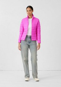 Giacca imbottita aderente di colore rosa acceso, con motivo trapuntato orizzontale, collo alto e chiusura con cerniera. Abbinata a jeans azzurri.