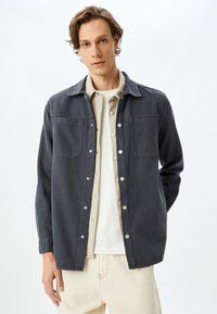 Chaqueta de un gris oscuro, texturizada, con dos bolsillos en el pecho, cierres de botón y un collar, llevada sobre una camisa de color claro y pantalones beige.