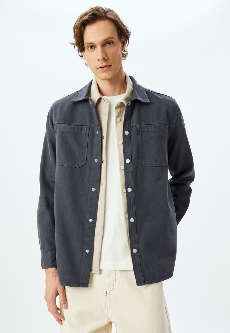 Chaqueta de un gris oscuro, texturizada, con dos bolsillos en el pecho, cierres de botón y un collar, llevada sobre una camisa de color claro y pantalones beige.