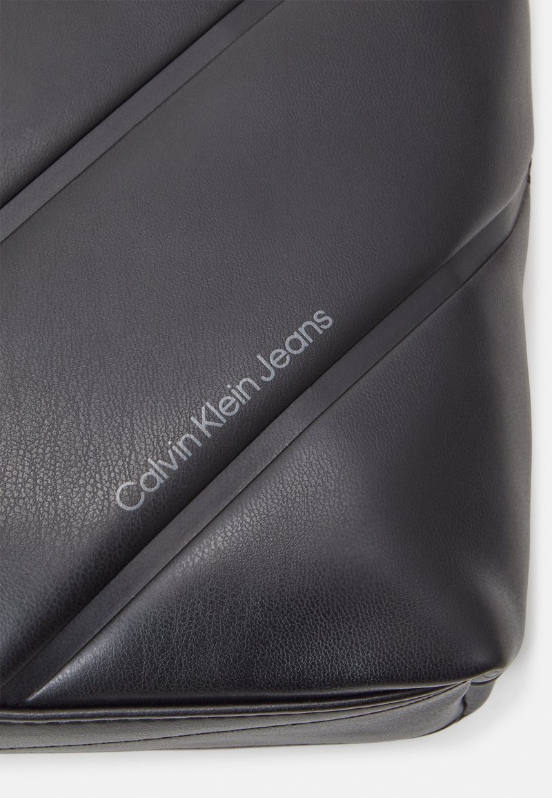 Black synthetic leather bag with diagonal seams, embossed "Calvin Klein Jeans" logo, smooth texture, and soft structure.