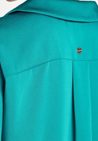 Turquoise satin blouse featuring a back pleat, wide collar, and a metallic logo accent near the neckline for added detail.