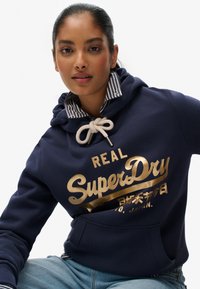 Donkerblauwe hoodie met gouden "SuperDry" logo, een trekkoord en gestreepte kraag, gemaakt van zachte stof met een voorkantzak.