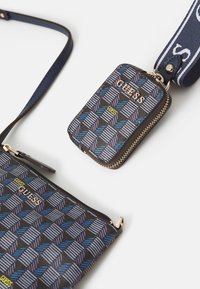 Borsa a tracolla e pochette piccola con cerniera, con motivo geometrico blu e viola, loghi Guess dorati e tracolla blu scuro su sfondo bianco.