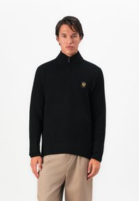 Homem a usar um suéter preto de malha com fecho, com um pequeno logótipo no peito e calças bege, de pé contra um fundo branco.