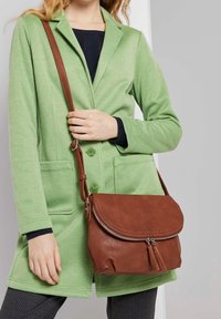 Brun axelväska i läder med rundad klaff, dragkedjestängning och tofsdetalj. Bärs över en grön texturerad kappa med fickdetaljer.