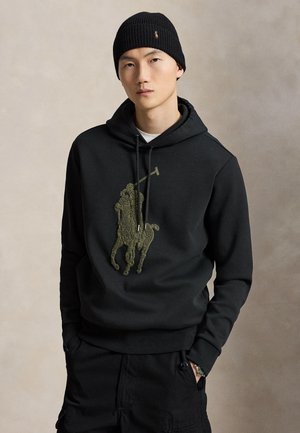 Jonge man met een zwarte beanie en zwarte hoodie met een groot getextureerd polospelerlogo, staand met handen in de zakken tegen een effen achtergrond.