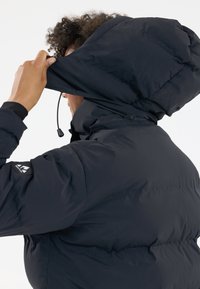 Person, der die Kapuze einer schwarzen gepolsterten Winterjacke mit sichtbar zip und einem Logo auf dem Ärmel vor einem schlichten Hintergrund anpasst.