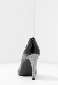 Zapato de tacón alto de charol negro con un tacón geométrico texturizado en gris, que presenta una punta redondeada y un acabado suave.