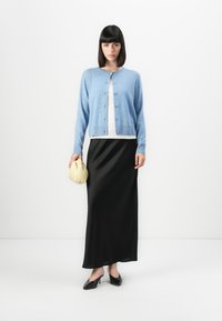 Lys blå cardigan, hvit topp, svart maxiskjørt, holder en gul clutch; glatt tekstur og enkel knappedesign. Svart spisse hæler.