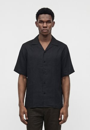Homme adulte aux cheveux courts portant une chemise noire à manches courtes boutonnée et un pantalon sombre, debout devant un fond clair uni.