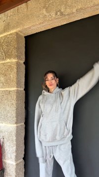 Pilka maikutė su kapom ir sportinės kelnės, pagamintos iš minkštos medžiagos; stovi priešais tamsias duris, matoma akmeninė arkos tekstūra.