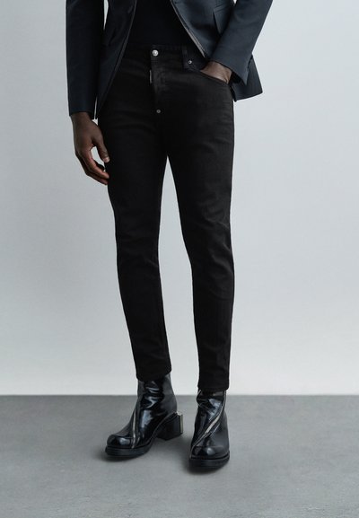Homme portant un jean noir slim, un blazer noir et des bottines en cuir noir avec des fermetures éclair à l'avant, debout sur un sol gris.