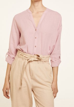 Kvinde iført en lyserød skjorte med knapper, ærmerne rullet op, og beige bukser med høj talje, bundet med et stofbælte.