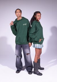 Sweatshirts à capuche vert foncé avec poches avant, en matière légère et avec un branding discret. Les modèles sont associés à des jeans déchirés et une jupe en denim, ainsi qu'à des bottes accentuées.