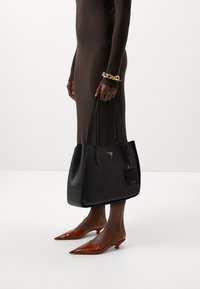 Czarna torba typu tote z wytłaczanym wzorem logo, podwójnymi uchwytami i małą metką. Modelka ma na sobie brązową sukienkę w prążki i brązowe buty na szpilce.