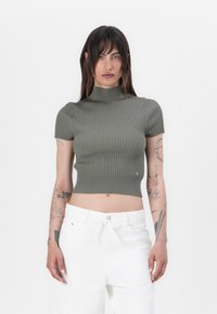 Olivgrünes, geripptes Cropped-Top mit hohem Kragen und kurzen Ärmeln, verfügt über ein dezentes Logo-Detail am Saum. Trägt man mit weißen, hoch taillierten Hosen.