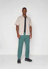 Άνδρας στέκεται φορώντας μπεζ κοντομάνικο πουκάμισο με κουμπιά πάνω από μαύρη μπλούζα, πράσινο παντελόνι και μαύρα αθλητικά παπούτσια, σε απλό φωτεινό φόντο.