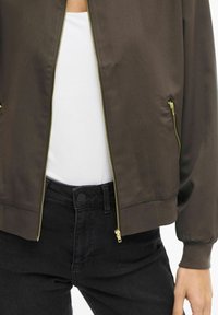 Brun bomberjakke med glidelås foran, sidelommer og ribbestrikkede mansjetter. Brukt over en hvit topp og kombinert med svarte jeans. Med blank finish.