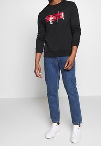 Uomo che indossa un felpa nera a maniche lunghe HUGO, jeans blu e sneakers bianche, in piedi contro uno sfondo grigio chiaro uniforme.