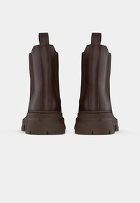 Een paar donkerbruin leren Chelsea boots met dikke profielzolen, gezien van de achterkant op een witte achtergrond.