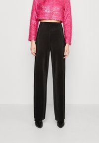 Persona vestida con pantalones negros de pierna ancha y cintura alta, junto con una blusa corta de manga larga rosa y peluda, de pie contra un fondo claro y sencillo.