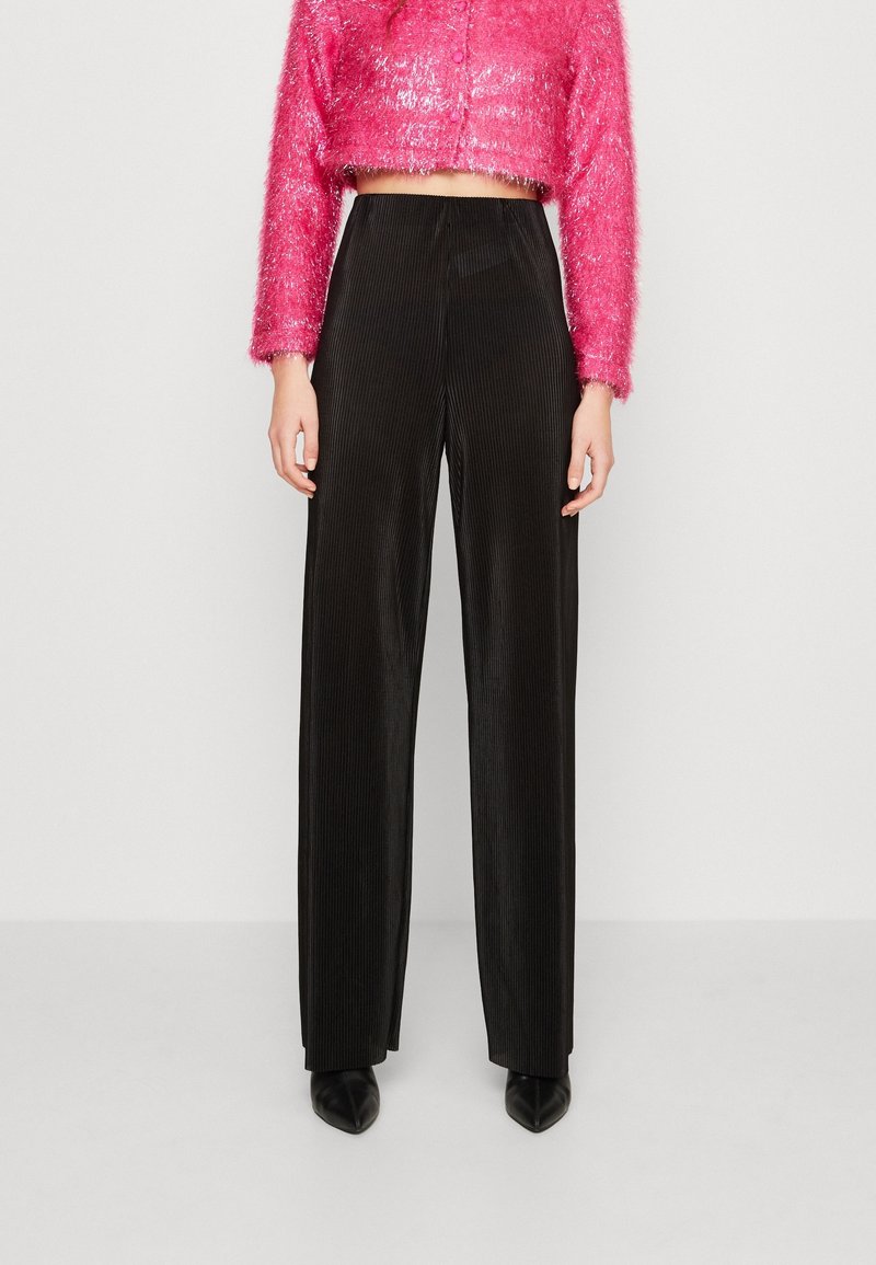 Persona vestida con pantalones negros de pierna ancha y cintura alta, junto con una blusa corta de manga larga rosa y peluda, de pie contra un fondo claro y sencillo.