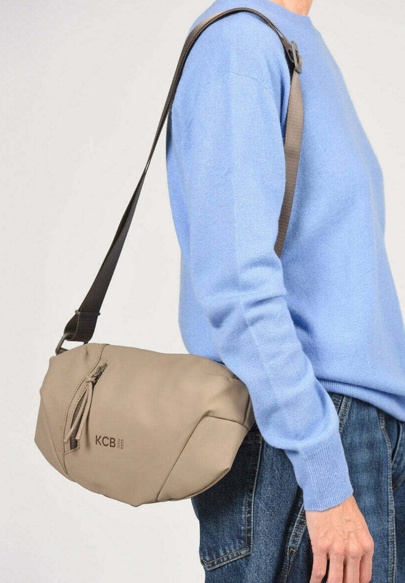 Beige schoudertas met ritssluiting en kwastje, gedragen over de schouder, gecombineerd met een lichtblauwe trui en spijkerbroek.