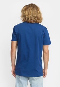 Camiseta de algodón azul con manga corta y cuello redondo, exhibida desde la parte de atrás, combinada con jeans de mezclilla claros. Textura suave.