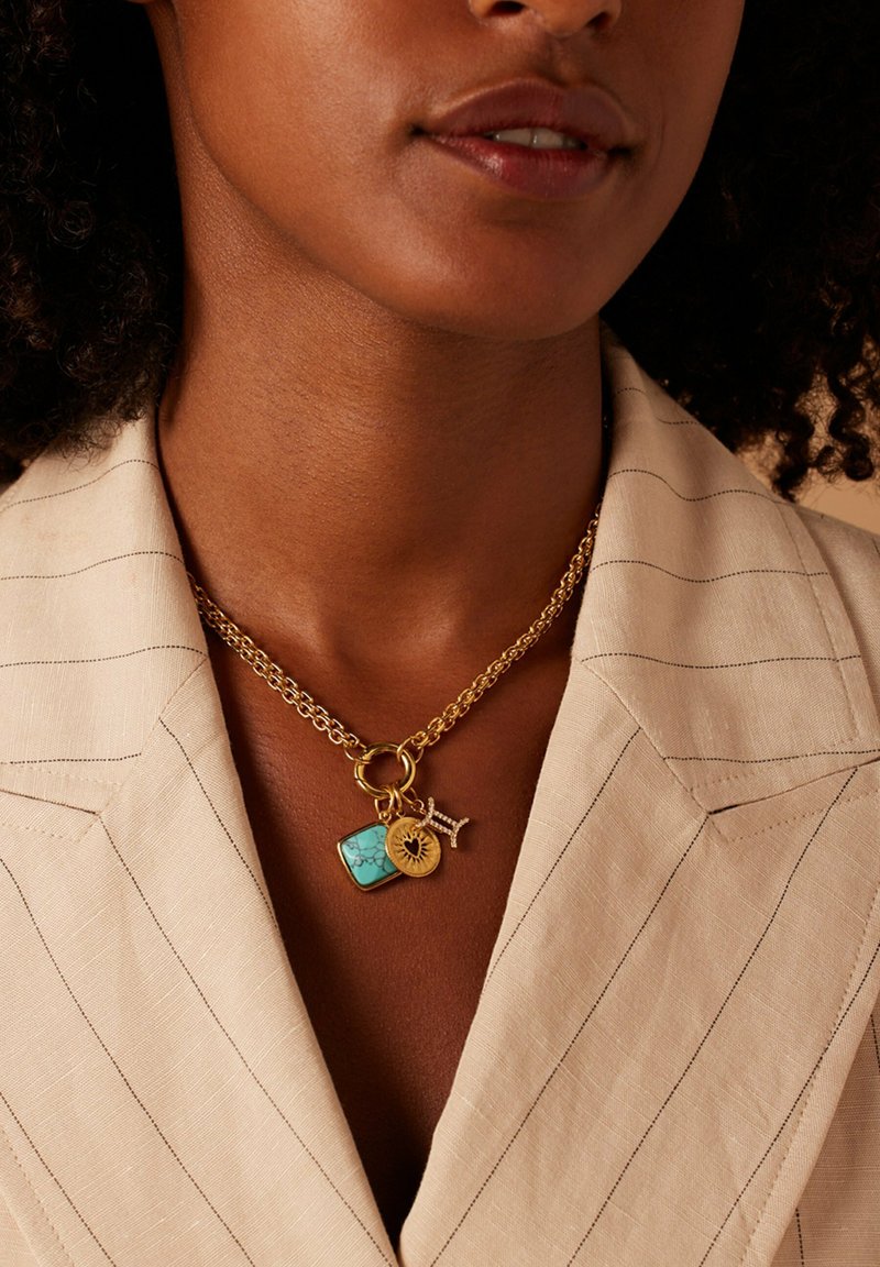 Gouden ketting met een turquoise rechthoekige hanger en bedels, gedragen over een lichtgekleurde, gestreepte blazer met een gesneden kraag.