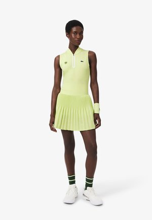 Mujer con vestido de tenis sin mangas color verde claro con cremallera frontal, muñequera a juego, calcetas a rayas verdes y zapatos deportivos blancos, posando con confianza.