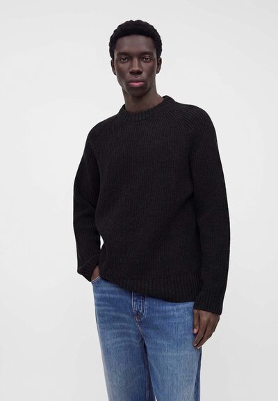 Jeune homme portant un pull en maille noir et un jean bleu, debout avec une main dans la poche devant un fond clair uni.