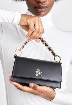 Kobieta w białym golfie trzymająca czarną torebkę z złotym łańcuszkowym uchwytem i złotym logo na przedniej klapie.