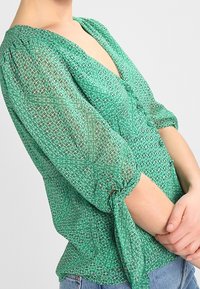 Blouse verte à motifs avec un col en V, des manches bouffantes et un détail noué aux poignets. Fabriquée en tissu léger et texturé.