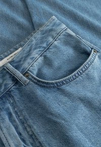 Το ανοιχτό μπλε τζιν ύφασμα διαθέτει ένα μπροστινό τσέπη με στρογγυλεμένο σχήμα, αντίθετες ραφές και μεταλλικό κουμπί κοντά στη μέση.