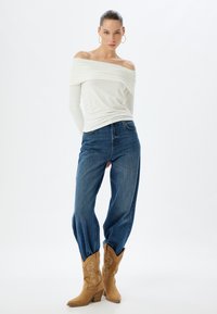 Witte off-the-shoulder blouse met geplooide textuur, gecombineerd met losse blauwe denim jeans en tan cowboylaarzen met decoratieve stiksels.