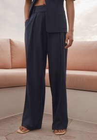 Marineblaue, taillenhohe, weit geschnittene Hose mit Falten, aus leichtem Stoff. Kombiniert mit beigen Sandalen, vor einem Hintergrund aus einem weichrosa Sofa.