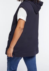 Henkilö, joka on pukeutunut mustaan hihattomaan huppariin valkoisen lyhythihaisen paidan ja sinisten farkkujen päälle, seisoo kyljellään yksinkertaista taustaa vasten.