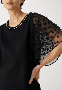 Top negro con escote redondo adornado con detalles metálicos. Mangas negras transparentes con lunares y volantes en capas. Textura de tela suave.