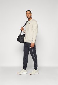 Sweatshirt bej deschis cu manșete ribbate, asociat cu pantaloni atletici bleu închis cu dungi laterale albe. Geantă de gimnastică neagră, adidași bej.