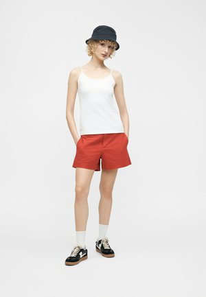 Γυναίκα με μαύρο καπέλο τύπου bucket, λευκό αμάνικο τοπ, κόκκινα σορτς, λευκές κάλτσες και μαύρα αθλητικά παπούτσια, στέκεται με τα χέρια στις τσέπες.