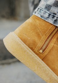 Botas de ante de color marrón con una cremallera lateral y una suela de goma texturizada. Detalles notables incluyen una cremallera dorada y el nombre de la marca en relieve en el costado.