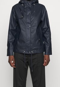 Mörkblå vattentät jacka med huva, tryckknappar och dragsko i midjan. Slät, matt yta med sidofickor och en lätt strukturerad design.