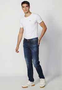 Fehér, rövid ujjú póló, kis narancssárga emblémával, sötétkék farmernadrággal, amelynek enyhe kopása van, fehér tornacipővel viselve.