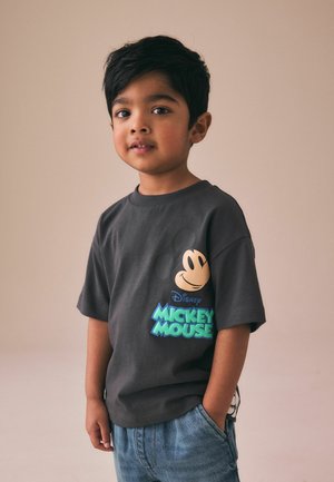 Young boy with short black hair wearing a dark gray Mickey Mouse T-shirt and blue jeans, standing against a plain background.