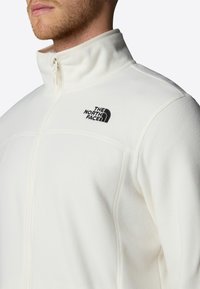 Chaqueta de forro polar blanca con cuello alto, que presenta una cremallera frontal y el logo negro de The North Face en el lado izquierdo del pecho. Textura suave y diseño minimalista.