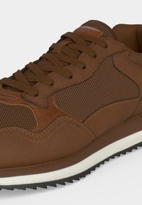 Baskets marron avec panneaux en mesh et cuir, lacets marron assortis, semelle blanche avec bande de roulement noire, montrées en gros plan.