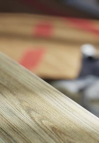 Gros plan d'une planche en bois clair avec un grain visible, arrière-plan flou montrant des planches empilées avec des marques rouges et un ruban à mesurer.