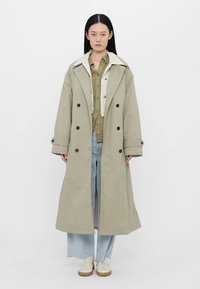 Long, double-breasted trench coat in light olive green with a cream collar, featuring large buttons and a relaxed silhouette.
