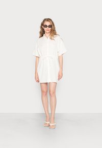 Vestido branco de camisa com mangas curtas, frente com botões e cintura ajustável. Estilizado com óculos de sol pretos e sandálias de salto brancas. Textura suave.