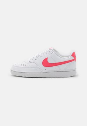 Białe niskie buty sportowe z różowym logo Nike, perforowanym noskiem i białą podeszwą na jednolitym jasnoszarym tle.
